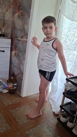 A young boy in a white tank top and black shorts is running through a home. He is barefoot. In the first few seconds, he is running past a doorway with a white frame and a patterned wall. He continues running into another room that appears to be a kitchen. Then he runs back into the first room and disappears through another doorway. The rooms have tile floors and one has a shoe rack with shoes on it. There is a bottle of soda and other items on a counter in the kitchen.