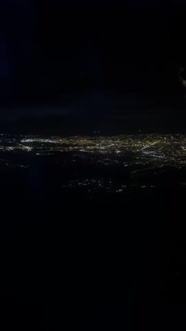 The video shows an aerial view of a city at night. Numerous lights illuminate the urban landscape, creating a sprawling network of yellow and white dots against the dark background. The lights appear to be concentrated in a central area, suggesting a densely populated city center. The perspective is from an elevated position, possibly from an airplane, given the slight angle and the limited visibility of the horizon. The overall impression is one of a vast, illuminated city from high above, with only the lights of human habitation visible in the darkness.