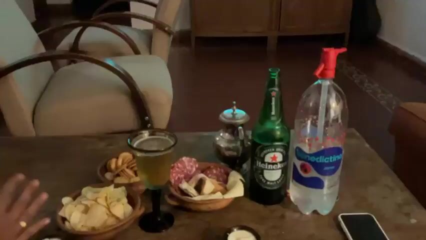 The video shows a living room with furniture and a television playing a soccer game. In the foreground, there is a table with a beer bottle, a spray bottle, and some food items. In the background, there is a fireplace, chairs, and a cabinet. The lighting is dim, suggesting it is nighttime or the room is not well-lit.