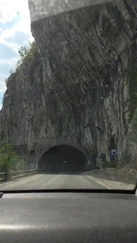 The video begins with a view from inside a vehicle looking out at the entrance to a tunnel carved into a rocky mountainside. The tunnel entrance is arched and constructed with stone blocks. As the vehicle enters the tunnel, the view shifts to the interior. The tunnel walls are rough and natural rock, with some sections showing rough-hewn stone. Reflective markers are visible along the sides of the road within the tunnel. The light at the end of the tunnel grows brighter as the vehicle progresses, indicating its exit. The video ends with the vehicle exiting the tunnel into bright daylight.