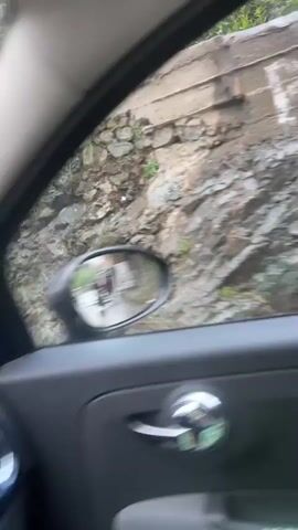 The video captures a car journey through a mountainous region in Asgaour, Morocco, characterized by wet weather.

The initial frames show a blurry waterfall cascading down a rocky, green hillside, viewed from the passenger side window of a moving vehicle. The scene quickly shifts to a view of the car's interior, then outside again, revealing a wet road and various structures.

Along the roadside, there are buildings with outdoor seating areas, some adorned with colorful accents like blue and pink, and surrounded by lush green trees. Concrete barriers line the road. In the car's side mirror, reflections show people walking and other vehicles on the wet road.

As the car continues, a vintage-style blue Mercedes-Benz van drives past on the opposite side of the road. In the background, majestic grey mountains loom under a cloudy sky, with some buildings featuring red roofs nestled among the greenery. The road remains visibly wet throughout, suggesting recent or ongoing rain. The car's side mirror also shows the arm of a person in a white t-shirt inside the car.