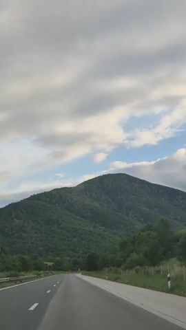 A video of a road trip. The camera is moving forward, suggesting the video was taken from a vehicle. The road is asphalt and has white dividing lines. On the left side of the road is a grassy shoulder, and beyond that, a green, tree-covered hill. On the right side of the road, there are more trees and a fence. In the distance, a large, green, tree-covered mountain rises against a cloudy sky. The sky is mostly covered in white and gray clouds, with some patches of blue visible. The lighting suggests it is either early morning or late afternoon. No people or specific actions are visible beyond the implied motion of travel.