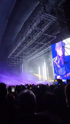 The video shows a concert crowd with people holding up their phones to record the stage.  The stage is brightly lit with white and purple lights. A large structure is visible above the stage, and a blue screen stands on the right. The crowd is silhouetted against the bright stage lights. The venue is an indoor space with a curved roof.  Some people in the crowd raise their index fingers up.