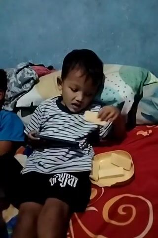 Two young boys are sitting on a bed with blankets. The boy on the right is wearing a striped shirt and black shorts. He is holding a phone and eating something from a yellow plate. The boy on the left is wearing a blue shirt and is also holding a phone. The room has a blue wall.
