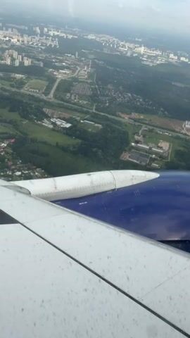 The video shows an aerial view from an airplane window. The wing of the plane is visible in the foreground, with the engine cover in blue and white. Below the wing, a sprawling city with numerous buildings and a network of roads can be seen amidst green trees and fields. The sky above is cloudy. The camera appears to be slightly tilted, giving a dynamic perspective of the landscape below.