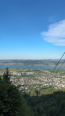 The video shows a view from a high vantage point overlooking a city and a large body of water under a clear blue sky with some white clouds. In the foreground, there are green trees and foliage, and several cables, likely from a cable car or zipline, stretch diagonally across the frame from the upper right to the lower left. The city appears to be densely populated with buildings of various sizes and colors, situated on the shore of the body of water, which extends into the distance. Beyond the city and the water, there is a hazy landscape of rolling hills and more distant settlements. The overall scene depicts a scenic, possibly urban or suburban, environment on a sunny day.