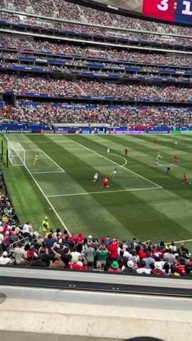 The video shows a soccer game from an elevated perspective. There are many spectators in the stands, wearing various colors. On the field, there are players in red and white uniforms. The action appears to be near the goal. A player in red is about to kick the ball, and another player in white is close by. The field is green with white lines, and there are advertisements around the stadium. The sky is visible above the stands.