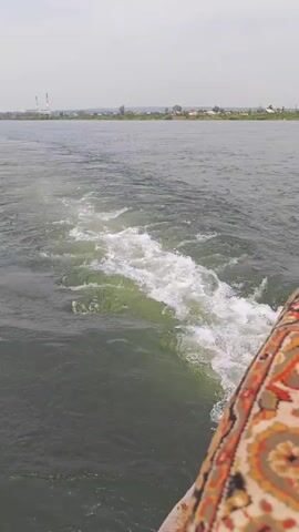 The video shows a wake from a boat moving through water, seen from the perspective of someone on the boat. The water is dark green and choppy, with white foam and churning visible in the wake. In the background, there is a shoreline with some buildings and trees, and a hazy sky. The right side of the frame shows a portion of the boat's interior, including a patterned fabric. The camera is tilted slightly downward, focusing on the water.