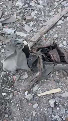 Okay, here's a detailed description of the scene shown in the video:

The video depicts a scene of heavy destruction and debris. The foreground consists of scattered rubble, including broken pieces of concrete, wood fragments, and crumpled sheets of metal. The ground is a mix of gray dirt and smaller debris. 

Moving towards the background, more large fragments of concrete are visible, along with twisted metal and other unidentifiable wreckage. The overall impression is one of a structure that has been heavily damaged or completely destroyed.

In the far background, green trees can be seen, suggesting this location is near or adjacent to some greenery, but the immediate environment is barren and filled with the remnants of devastation. There are no people visible in the video.