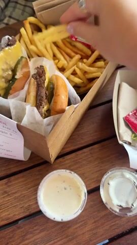 A hand is seen dipping french fries into a small container of sauce. The french fries are golden yellow and appear crispy. The sauce is light-colored and appears creamy. In the background, there are two burgers and a box of french fries. The scene is set on a wooden table.