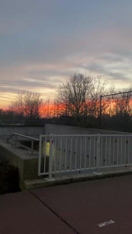 The video shows a concrete structure with a white metal fence in front of it, likely a pedestrian underpass, against a sky with a colorful sunset in the background. The sky has hues of orange, red, and gray, contrasting with the silhouetted trees and power lines. The camera is positioned on a paved surface, possibly a sidewalk or path, and it moves slightly during the recording. There are no people visible in the frame. The environment appears to be suburban, with elements of both natural and man-made structures.
