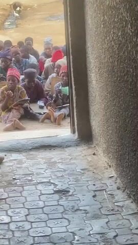 A group of children, most wearing head coverings, are seated on the ground in what appears to be an open-air or rudimentary classroom setting. The children are facing forward, engaged in an activity, with some holding books or papers. The ground is dusty and uneven. The foreground shows a textured, wet tiled surface and a rough wall. The perspective is from an entryway looking into the space where the children are gathered.