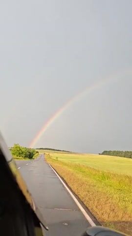 The video shows a road winding through a green landscape under a cloudy sky. A rainbow arches across the sky. The perspective is from inside a vehicle, with the side of the vehicle visible in the foreground. There are a few trees on the left side of the road and fields of grass and crops on both sides. In the distance, a car is driving away from the camera. The road appears wet, suggesting it may have recently rained.