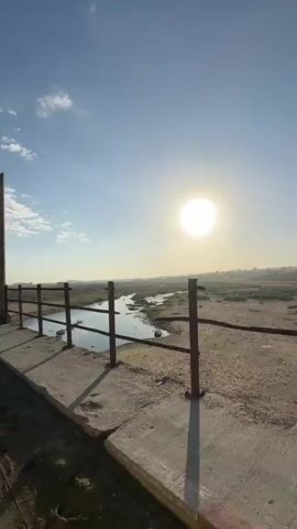 The video shows a slow pan across a body of water with a bright sun in the sky. The water appears to be shallow and dirty, with debris floating in it. The surroundings are dry and barren, with some sparse vegetation. There is a concrete structure with a metal railing in the foreground, which suggests the viewer is on a bridge or walkway. The overall impression is one of a desolate and possibly polluted landscape.
