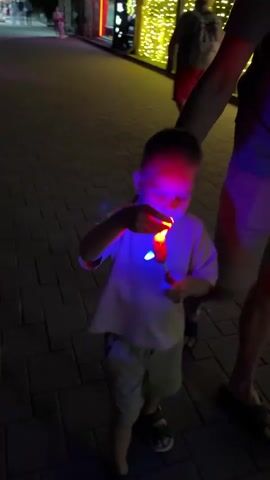 In this short video, a young boy is the central subject, holding a colorful, light-up toy that appears to be a spinning top or pinwheel. The toy emits bright red, blue, and green lights, casting a vibrant glow on the boy's face and hands. He is dressed in a light-colored t-shirt and shorts, and he's wearing sandals.

The scene appears to be outdoors at dusk or night, as indicated by the low light conditions and the illuminated string lights visible in the background. The ground is paved with interlocking bricks, suggesting a walkway or public space. An adult's arm and shoulder are visible on the right side of the frame, indicating their presence and possibly support for the child. Another person, mostly obscured, is seen in the background, also in the low light.

The boy seems captivated by the toy, his gaze focused intently on it as he manipulates it. The overall mood is one of simple childhood wonder and enjoyment, amplified by the dazzling lights of the toy and the festive atmosphere suggested by the background lights. The setting, with its brick paving and decorative lights, hints at a recreational area, possibly a boardwalk or park during an evening event. There is no visible text in the video.