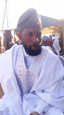 The video features two men wearing traditional Nigerian clothing, specifically a white robe with intricate geometric patterns and brown caps. The first man, with a full beard, stands and looks directly at the camera. He appears to be speaking, though the audio is not described. The camera then pans slightly to the side, showing the man sitting, and he smiles at the camera. The setting appears to be outdoors, possibly a gathering or event, with a temporary structure, chairs, and other people visible in the background. There is a sense of community and possibly celebration.
