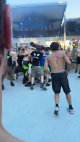 A group of young men are gathered in a circle on a paved outdoor area. Some are shirtless, others are wearing t-shirts and shorts. One man is being lifted by others, and several people are embracing. The setting appears to be a music festival or outdoor event, with a stage structure and lighting visible in the background. The overall atmosphere seems to be one of excitement and camaraderie.