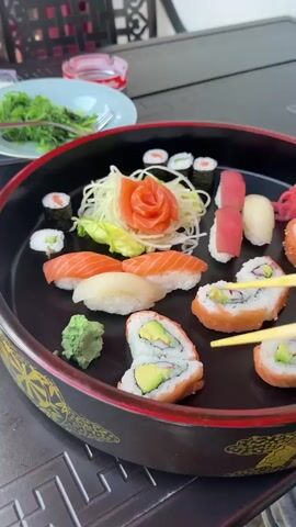 Here is a description of the video.

The video shows a person using chopsticks to pick up a piece of sushi from a black platter. The platter contains various types of sushi, including maki rolls, nigiri with salmon and white fish, California rolls with avocado and salmon, wasabi, and shredded radish. In the background, there is a plate of seaweed salad and an ashtray on a table. The person appears to be outdoors, as the background shows a patio setting.