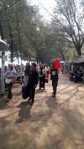 A group of people are walking along a sandy path lined with trees. A man in the foreground is carrying a large red bucket on his shoulder. To the left, there are motorcycles parked and a woman holding a child. On the right, there are small shops and stalls. The weather appears to be sunny.