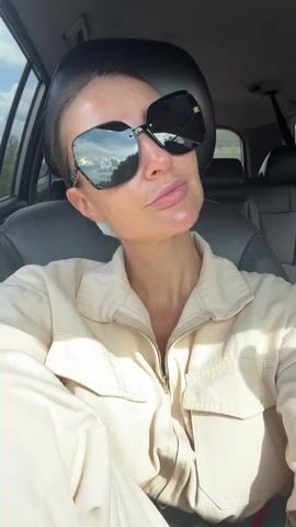A woman wearing large sunglasses and a beige jumpsuit sits in the driver's seat of a car. She looks towards the camera and smiles slightly. The car appears to be parked or stopped, as indicated by the static shot of the woman. The background shows the interior of the car and a view through the passenger window, which reveals trees and a blue sky with clouds. In the latter half of the clip, the view through the passenger window changes, showing trees, a dirt road, and a railroad crossing gate. The camera is held at an angle, suggesting it is a selfie taken from the driver's perspective.