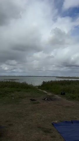 The video displays an outdoor scene near a body of water. The foreground shows a patch of dry grass with some scattered debris, including what appears to be remnants of a campfire and a blue tarp. A path leads from this area towards the water. The middle ground is dominated by tall, green grass and reeds lining the water's edge. The water itself is visible as a large expanse under a cloudy sky. In the background, there are faint landmasses or islands in the distance. To the right of the frame, a small green tent is partially visible, as is a white portable high chair with some items on it. There are no visible people in the video. The lighting suggests it is daytime, likely overcast.