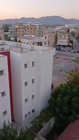 The video pans across a cityscape at dusk, focusing on several multi-story buildings. The foreground features the rooftops of red-tiled houses, some with solar panels. A white building with small square windows and a red accent stripe is prominent in the center. Behind it, more buildings of varying heights and architectural styles are visible, including some with balconies. Trees and vegetation are scattered throughout the urban landscape. In the distance, mountains form a backdrop against the pale evening sky. Cars are visible on streets, and there are signs of construction or storage in some areas. The overall impression is of a residential or mixed-use urban environment.