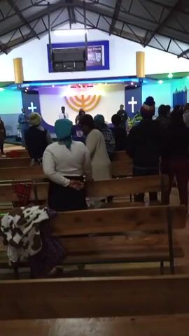 Certainly! Here's a description of the video:

The video shows a gathering of people inside what appears to be a church. Most individuals are standing in the pews, facing a raised stage area. On the stage, several individuals are positioned behind a lectern. The stage is decorated with two white crosses and a golden menorah. A screen displaying the words "City Church Witness" hangs above the stage. The interior of the building has a metal roof structure and is lit with colored lights. The audio suggests a religious service is in progress.
