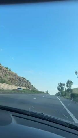 The video shows a view from inside a car driving on a highway. The sky is clear and blue. On the left side of the road, there is a rocky hill with some vegetation. On the right side of the road, there are trees and green hills. There are speed limit signs visible on both sides of the road, indicating a speed limit of 60. The car's dashboard is partially visible at the bottom of the frame, with a Mercedes-Benz logo. The road has white lines marking the lanes.