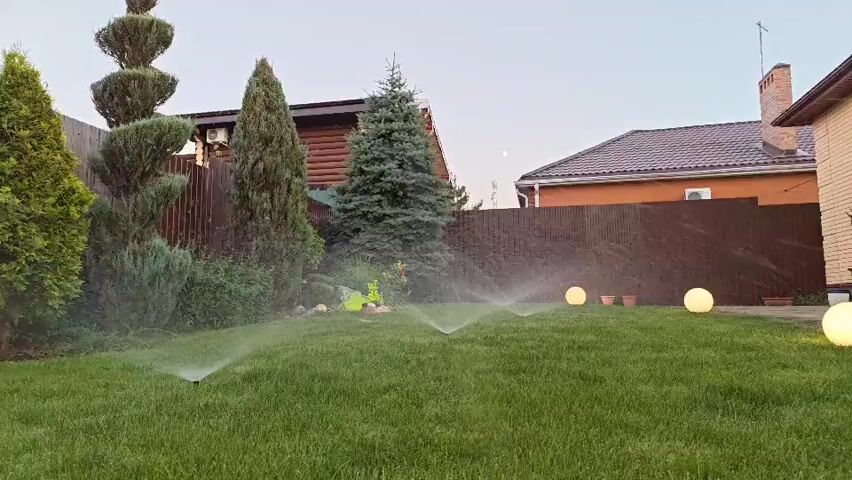 The video shows a backyard lawn with sprinklers watering the grass. The scene is set at dusk, with soft lighting from spherical outdoor lamps. Several trees and bushes are visible, along with a wooden fence and parts of two houses. There are no people or animals in the video, and the sprinklers appear to be the only moving objects, aside from the water they are spraying.