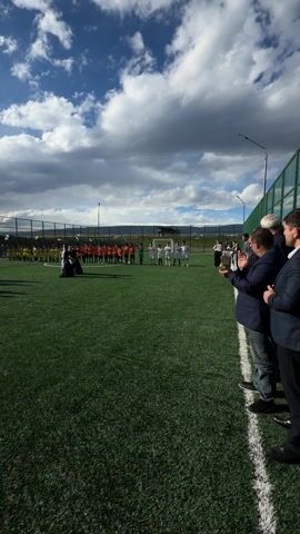On a sunny day, a soccer field is the scene of a unique event. Two teams, one in yellow and one in orange, stand facing each other on the grass, while six people in dark robes perform some sort of ritual or dance. The people in robes have their arms raised and are kneeling, creating a somber yet intriguing spectacle. Some spectators stand along the sidelines, clapping, while the sky above is filled with fluffy clouds. The field has a green surface, and the whole scene is a blend of sport and possibly cultural expression.