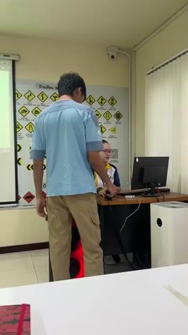 The video shows an indoor scene, likely a classroom or training room, with two individuals present. A young man with black hair, wearing a light blue shirt and khaki pants, walks from left to right in front of the camera. To his right, a woman is seated at a desk wearing glasses and a blue and yellow collared shirt. She is looking at a computer screen, and there is a sign behind her with various yellow warning symbols. A projector screen displays some information, and a speaker is located next to the desk. The room is well-lit and appears to be clean and organized.