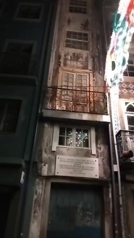The video shows a nighttime view of a narrow street with tall, old buildings. The building on the right is weathered and shows signs of age, with peeling paint. It has several windows, some with balconies. The window on the second floor from the bottom has Christmas lights visible inside. Below this window, a plaque with text is mounted on the wall. The building on the left is painted a teal color and appears to be newer or better maintained, also featuring windows and balconies. A colorful, possibly decorative, light string is visible at the top right of the frame. The street is dark, and the focus is on the facades of the buildings.