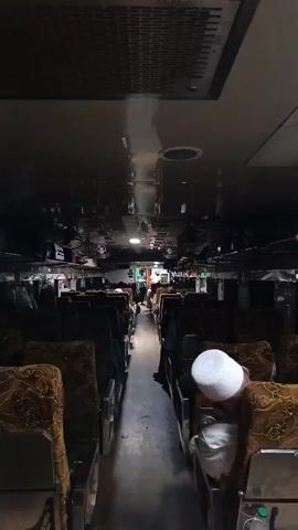 The video shows the interior of a train carriage. Rows of seats with brown patterned upholstery are visible on both sides of a central aisle. The ceiling is dark and reflective, with air vents and lights. Several passengers are seated, some appearing to be sleeping. One person in the foreground is wearing a white cap. The aisle appears to be empty in the foreground but shows a few figures further down the carriage. The lighting is dim, suggesting it may be nighttime or an indoor environment.
