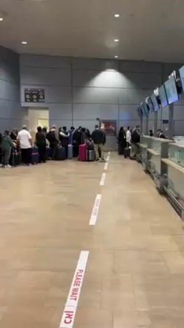 The video shows a long line of people waiting to check in at an airport. They are standing in a queue marked by floor stickers that say "PLEASE WAIT HERE." Most people have luggage with them, including suitcases and carry-on bags. The check-in counters are visible to the right of the queue. The environment appears to be an airport terminal with a tiled floor and a high ceiling.
