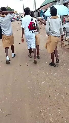 A group of at least five young people, appearing to be students in uniforms, walk along a dirt road. The uniforms consist of light-colored shirts and tan skirts or shorts. Some are carrying bags, and one person has a red backpack. The road is lined with what appears to be market stalls or small shops, with some structures covered by corrugated metal roofs. A few vehicles are visible in the background. The lighting suggests it is daytime.