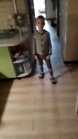 A child walks toward the camera in a narrow hallway with wood-patterned linoleum flooring. To the left is a kitchen area with a light green cabinet and a wire rack holding items. To the right is a closed white door and a small wooden table. The hallway leads to a sunlit room in the distance. The child is wearing a long-sleeved patterned shirt, brown pants, and black shoes.