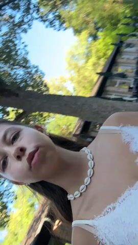 The video begins with a young woman in a white lace top and a seashell necklace looking at the camera. She is outside with trees and a building in the background. A sign that reads "Bad Orb Indoor-Gradierwerk & Cafe im Kurpark" is visible behind her. She smiles and turns her head. The camera then pans to show more of the environment, including the building and a paved walkway. The video ends with a blurred shot of the building.