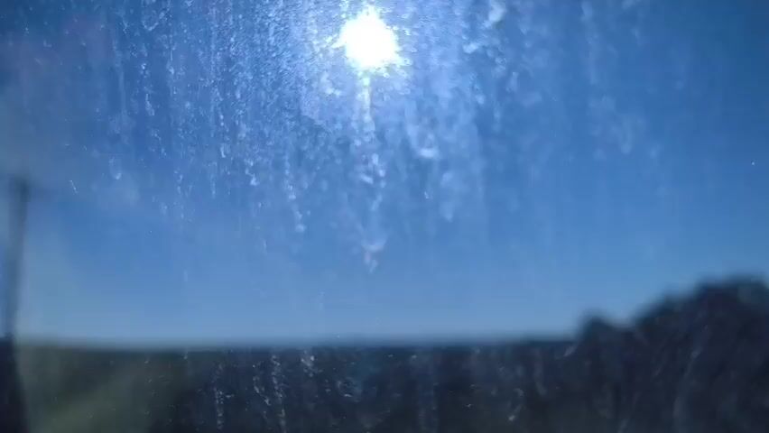 A close-up shot of a dirty window with the sun shining brightly through it. The sky is a clear blue, and a dark silhouette of a landscape can be seen in the distance. The camera appears to be moving slightly, possibly indicating that the shot was taken from a moving vehicle.