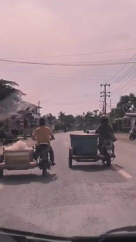 Two men on motorcycles with sidecars are traveling down a road. The motorcycle in the foreground has a yellow shirt-wearing man driving and the sidecar is carrying a light-colored box wrapped in plastic. The motorcycle in the background has a person wearing a dark shirt and helmet driving, with a large blue and green box in the sidecar. The road appears to be paved and lined with buildings on the left side. Overhead power lines are visible. The sky is overcast.