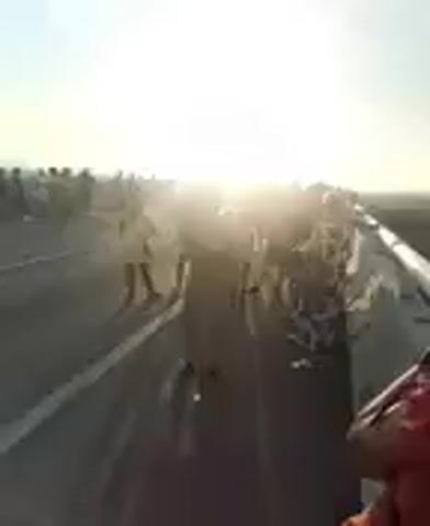 The video opens with a wide shot of a highway at sunset. A crowd of people are gathered on the highway, some standing, some walking. The sun is bright and low in the sky, casting a warm glow over the scene. The people appear to be facing the sun, and some are holding up phones or cameras, suggesting they are taking pictures or videos. There are cars or other vehicles on the side of the highway. The overall impression is one of a peaceful gathering, possibly to witness the sunset.