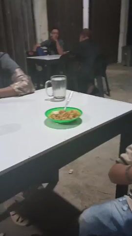 This media file captures a moment at an outdoor food stall or restaurant in Manyang Cut, Indonesia. The scene is dimly lit, suggesting it is nighttime or late evening. The foreground features a young child, likely a boy, actively eating something. He is wearing a light brown polo shirt and blue jeans, and has a determined expression as he brings food to his mouth with his hands. His left wrist sports a simple bracelet.

To the left of the child, a white table is visible, holding a green bowl with noodles and a fork, and a glass of water. Further back, other tables and chairs are arranged, with several other individuals present. One person, wearing a hijab, is seated at a table in the middle ground, engrossed in their smartphone. In the background, other patrons are seated at tables, some appearing to be smoking. The overall atmosphere seems casual, a typical scene at a local eatery. The environment suggests an open-air setting, possibly with a corrugated metal roof visible in the background. No visible text is present in the media.