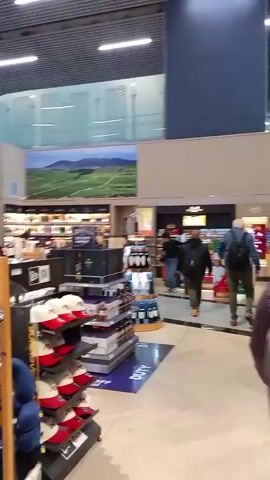The video shows the interior of a duty-free shop at an airport. In the foreground, there are displays of hats and other merchandise. In the background, there are shelves stocked with bottles of alcohol. Several people are visible browsing the store. One person is seen pulling a suitcase. The store appears to be well-lit and modern.