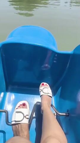 The video shows a first-person perspective from a blue paddle boat on a body of water. The viewer's feet, wearing white sandals with gold buckles and painted toenails, are visible at the bottom of the frame, resting on the boat's pedals. The water is calm and has a greenish hue. In the background, there is a shoreline with lush green trees, including weeping willows, and some buildings. A power pylon is also visible in the distance. The sky is clear and blue, suggesting a sunny day. The overall mood appears relaxed and leisurely, indicative of a pleasant outing on the water. No visible text is present in the video. The scene is likely a park or recreational area in Vahdat, Tajikistan.