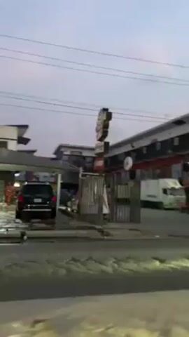 The video shows a street scene in a city. A building with multiple businesses lines the right side of the street. Various signs and billboards are visible on the building. People walk on the sidewalk in front of the building. Cars and trucks drive on the street. The sky is overcast and the lighting suggests it might be early morning or late evening. Overhead power lines run across the top of the frame.