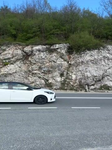 The video captures a scenic drive on a winding mountain road in Očinići, Montenegro, during the daytime under a clear blue sky with scattered white clouds. The road is asphalt with clear white lane markings, flanked by a steep, rocky mountain face covered in green trees and bushes on one side, and a gravel shoulder leading to a vegetated slope on the other.

Multiple cars are seen driving on the road. Initially, a white hatchback with a black roof is seen driving away from the camera. As the camera pans, a silver BMW 3 Series Touring (E91) drives past, followed by other vehicles in the distance navigating a curve. A blue sedan, possibly a taxi, is also seen driving in the opposite direction.

The overall scene depicts a peaceful journey through a natural, mountainous landscape, with cars intermittently passing by. A metal guardrail is visible along a section of the road. The weather appears pleasant, with bright sunshine highlighting the lush greenery and rocky textures.