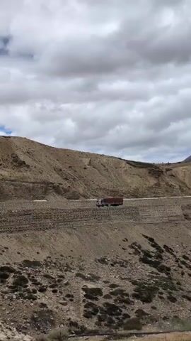 A wide shot shows a red and blue truck driving on a paved road winding through a barren, mountainous landscape. The road is built on a series of terraced slopes, with some vegetation visible on the lower slopes. The sky is partly cloudy with white cumulus clouds. The terrain is dry and rocky, with sparse scrub vegetation. The truck is moving from left to right across the frame.