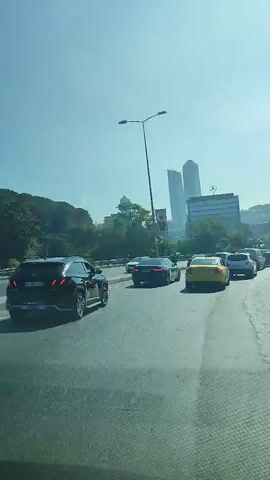 The video shows a view from inside a vehicle driving in heavy traffic on a sunny day. The road is asphalt and appears to be a multi-lane highway or a large city street. On the left side of the road, there are several cars, including a black sedan and a yellow taxi. Further in the distance, there are tall modern buildings, some of which appear to be skyscrapers. A lamppost is visible on the left. The road curves to the right, and on the right side, there is a steep, tree-covered embankment. More cars are visible ahead, indicating a traffic jam. The sky is clear and blue. The video is shot from a low angle, suggesting the camera is mounted inside a car.