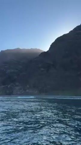 The video shows a dramatic landscape, likely a coastline, dominated by a rugged, dark mountain range. The sun is positioned high behind the mountains, creating a backlighting effect that highlights the rough texture and contours of the peaks. The water in the foreground is a deep blue, suggesting a clear, deep body of water. Small waves lap at the shoreline, indicating a gentle sea. The sky is a clear, pale blue, suggesting a serene, possibly early morning or late afternoon setting.