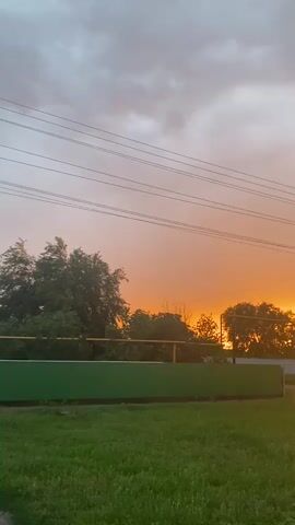 This is a vertical video of a rural landscape at sunset. The scene is dominated by a dramatic sky, with hues of orange, pink, and gray clouds. The sun is setting behind a line of trees, casting a warm glow.

In the foreground, there is a grassy field and a green fence. Behind the fence, power lines stretch across the frame. The trees are silhouetted against the bright sky. In the distance, houses and a utility pole are visible.

The time of day is sunset, as indicated by the warm colors in the sky. The weather appears to be partly cloudy. The location cues suggest a rural area in Taganrogskii, Russia. The overall mood is peaceful and serene. There is no visible text in the video.