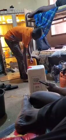 In a messy interior space, a person wearing an orange shirt and patterned pants bends over a white plastic jug with the words "HSP HEAVY DUTY SAE 40 API/SF 25 litres." The person holds a yellow container, possibly pouring something into the jug. A second person sits in the foreground, partially visible, holding a phone. One of their bare feet is prominent in the lower left. The room contains a bunk bed, a shelf filled with items, a gas cylinder, and various other objects contributing to the cluttered appearance.
